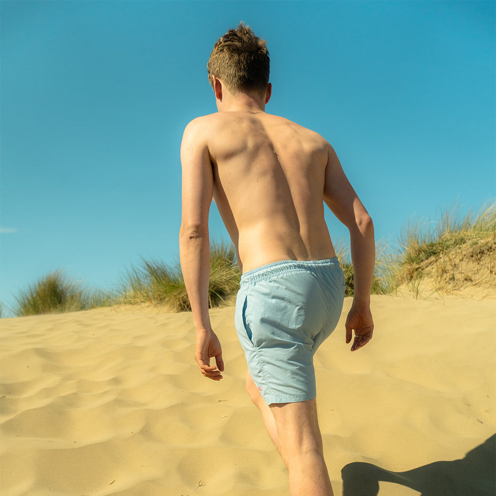 Man wearing pale blue swim shorts climbing sand dunes with clear blue sky