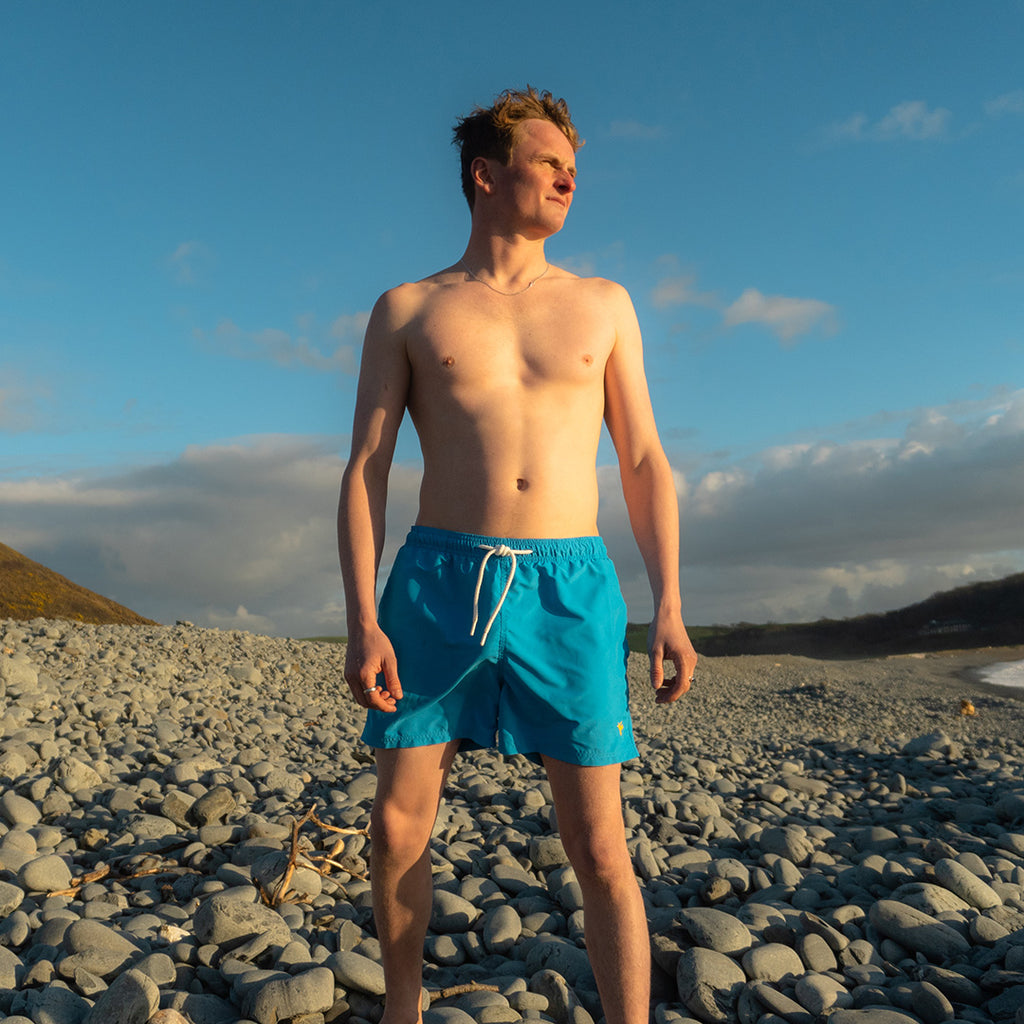 Man in blue swim shorts standing on a rocky beach with a blue sky and clouds in the background.