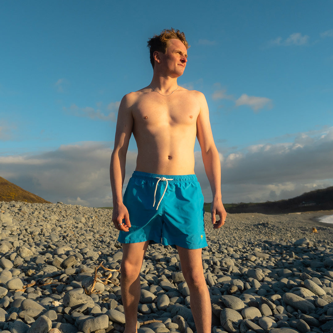 Man in blue swim shorts standing on a rocky beach with a blue sky and clouds in the background.