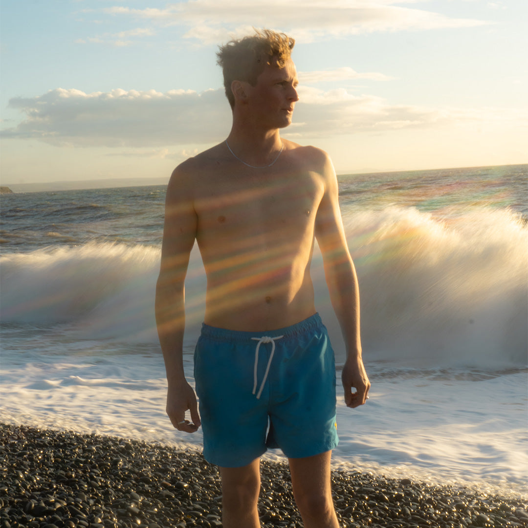 Men wearing blue Rolf Skeldon sustainable SEAQUAL swim shorts standing on a beach with waves and sunset.