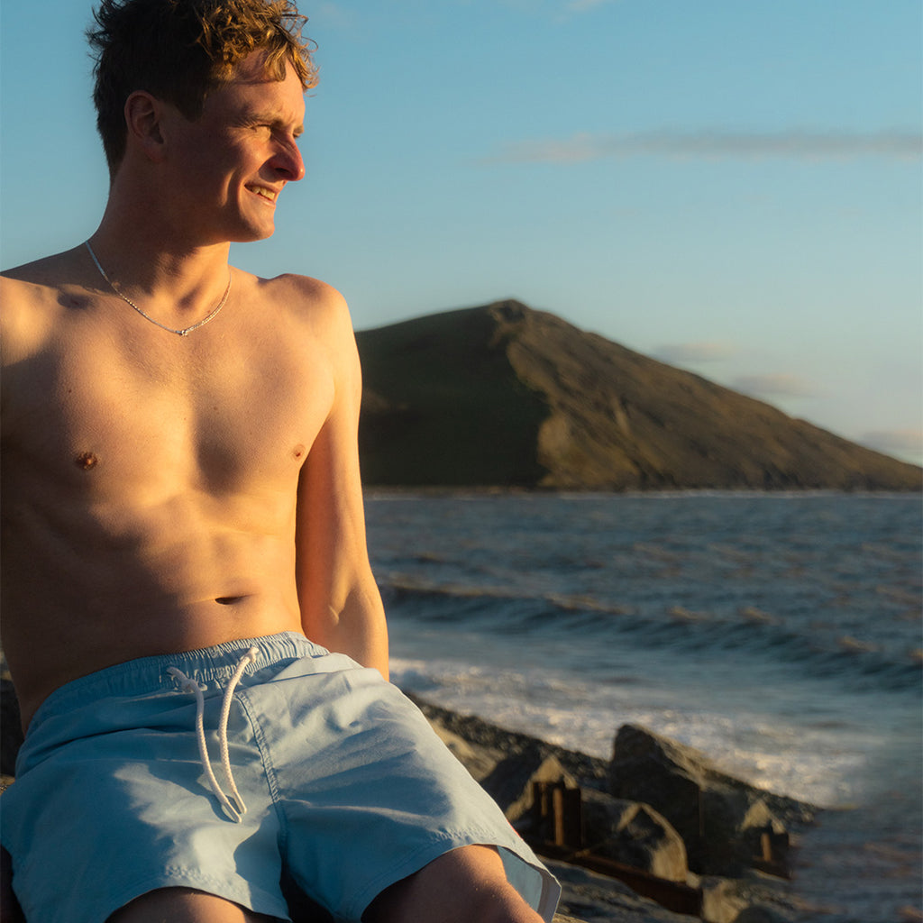 Men wearing pale blue Rolf Skeldon sustainable SEAQUAL swim shorts sitting on rocks by the ocean with a mountain in the background