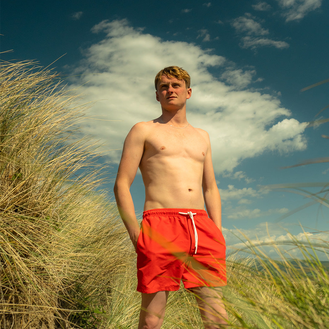 Man in red swim shorts standing in tall grass with a blue sky and clouds in the background