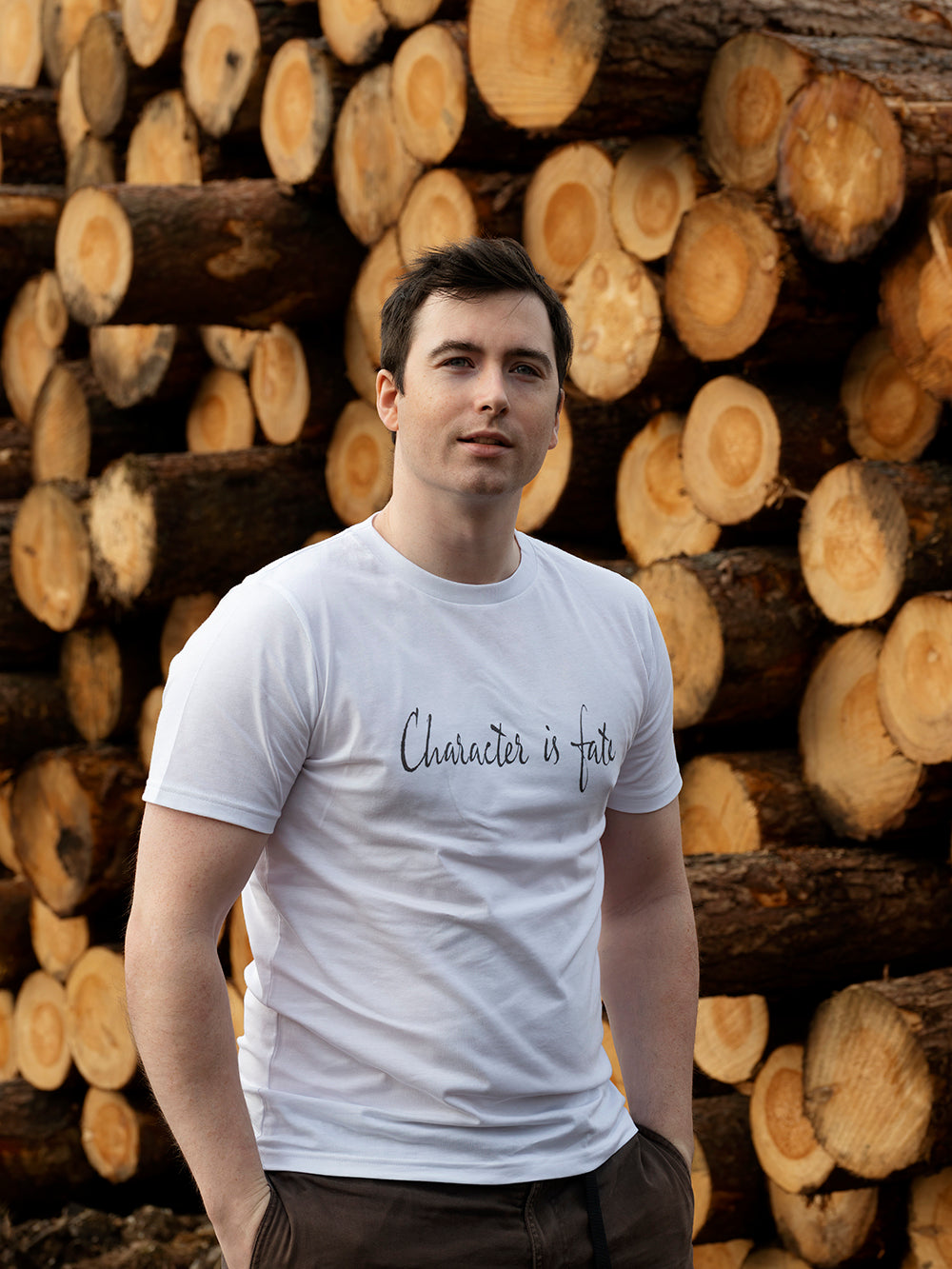 Man wearing a white t-shirt with text standing in front of stacked logs