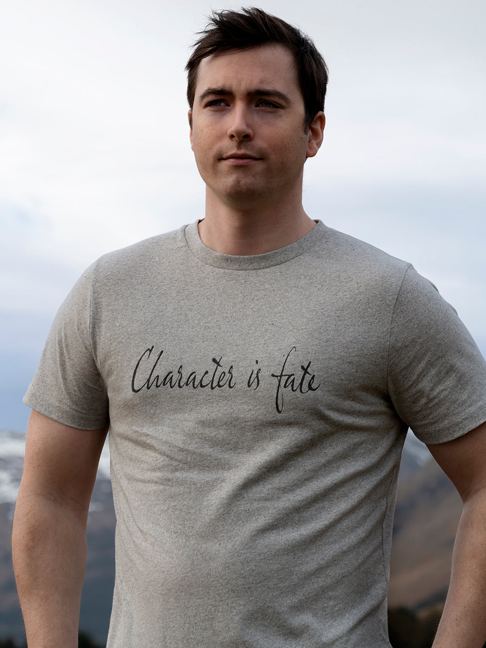 Man wearing a grey t-shirt with 'Character is fate' text against a cloudy sky.